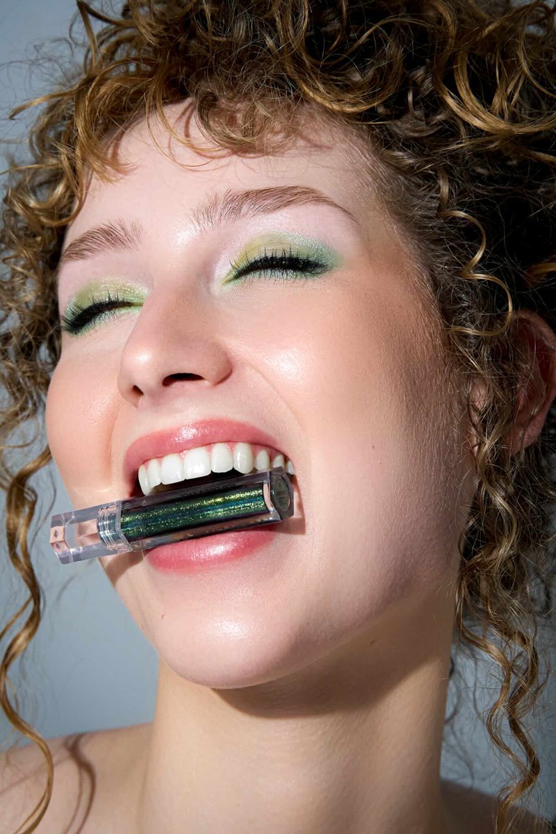Model mit grünem Lidschatten, lachend mit Lidschattentube im Mund